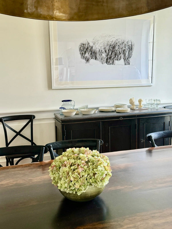A dome of green hydrangea in a low olive-glazed footed bowl, centred on a dark wood dining table with black chairs.