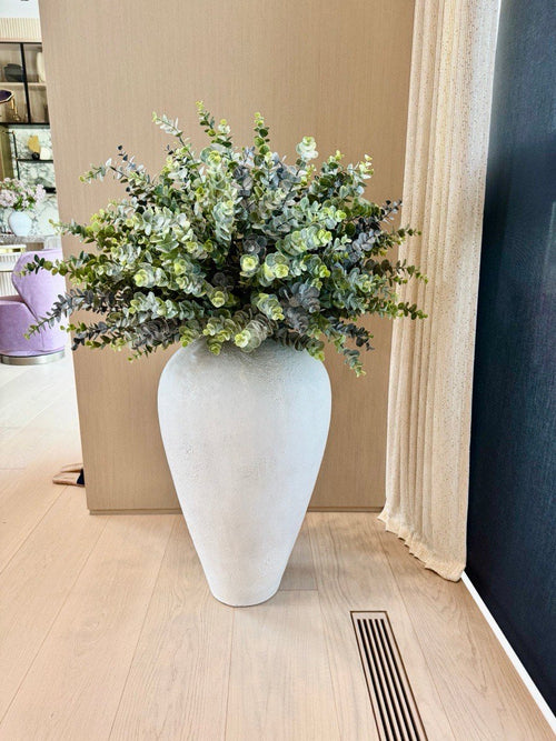 Eucalyptus and green foliage in a large white textured floor vase, standing on a light wood floor beside a curtain.