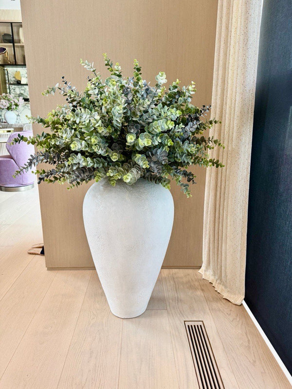 Eucalyptus and green foliage in a large white textured floor vase, standing on a light wood floor beside a curtain.