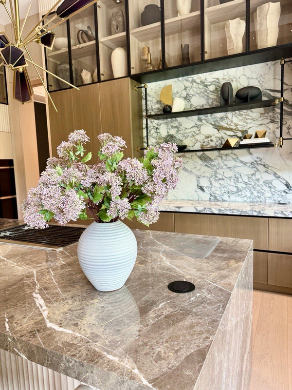 Lilac blooms in a ribbed white ceramic vase on a marble kitchen island, with a marble backsplash and wood cabinetry behind.