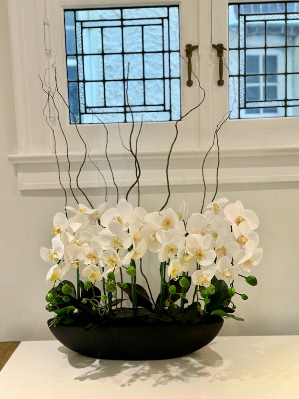 Cream white orchids with tall curly willow branches in a long black bowl, on a console in front of a leaded-glass window.