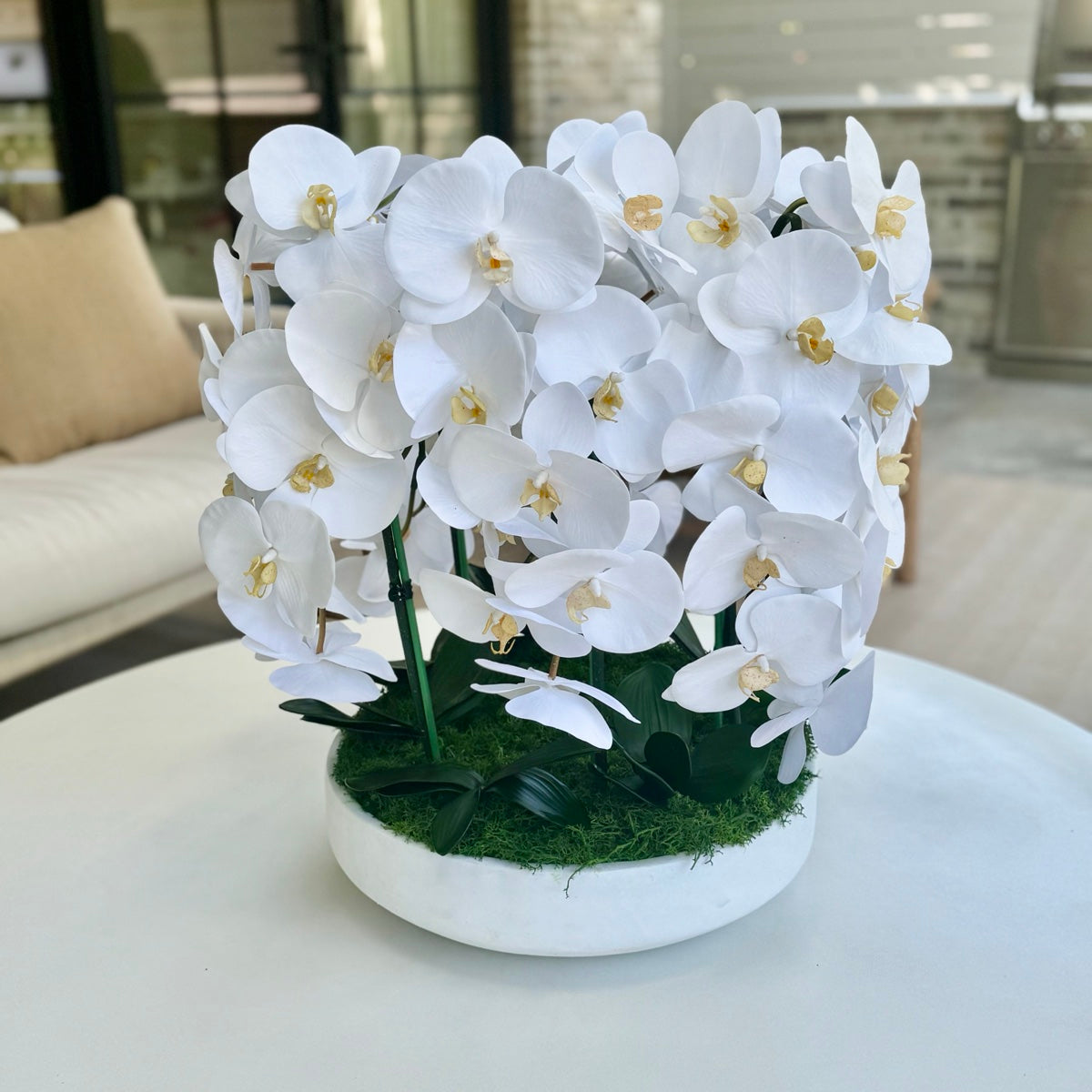 A dense dome of white orchids in a low white ceramic bowl with moss at the base, on a round table in a living room.