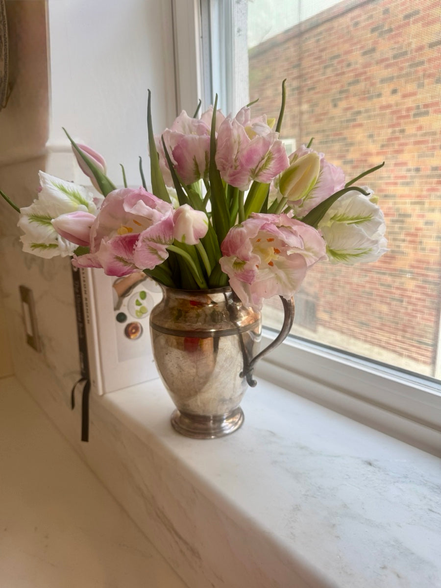 Pink and white tulips in a silver pitcher on a marble windowsill, with a brick building visible through the window.