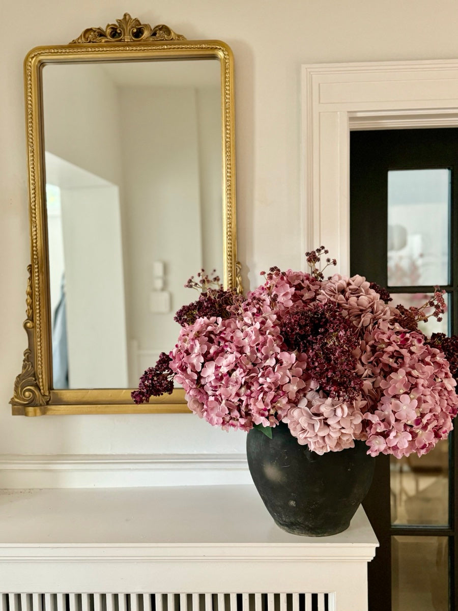 Pink hydrangea with deep burgundy stems in a dark matte pot, sitting on a white mantel below a gold-framed mirror.