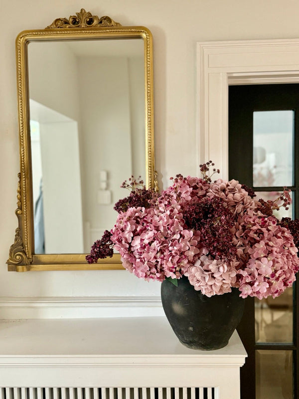 Pink hydrangea with deep burgundy stems in a dark matte pot, sitting on a white mantel below a gold-framed mirror.