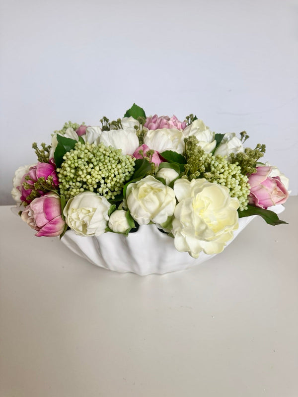 Pink and cream peonies with green blooms in a white scalloped bowl, photographed against a plain pale background.