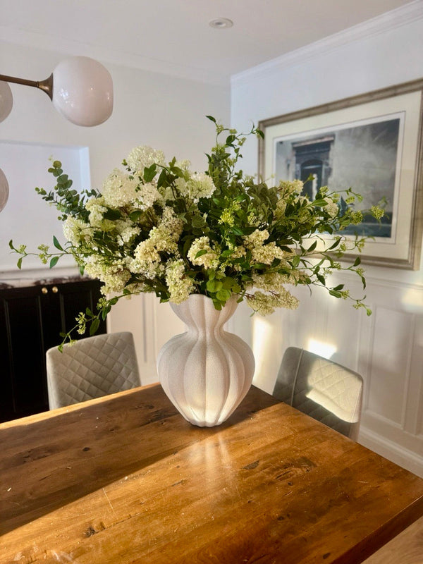 Cream white hydrangea and trailing greenery in a fluted cream ceramic vase, on a wood dining table with framed art behind it.